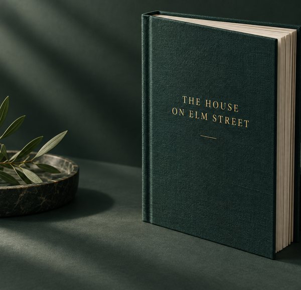 A deep green clothbound hardcover with a gold foil title stands on a dark table beside an olive sprig in a stone dish.