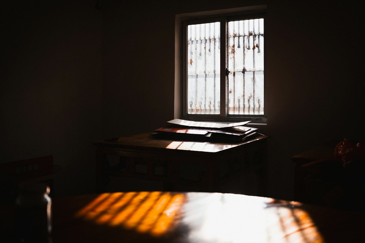 A quiet room with a desk beneath a barred window, sunlight falling across the table.