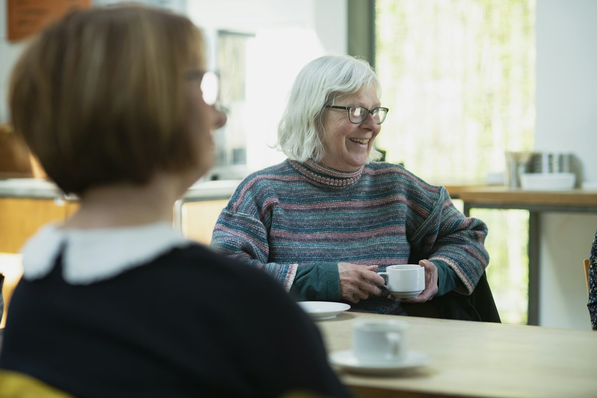 An older woman with a cup of tea smiles as she talks with a younger woman across a table.