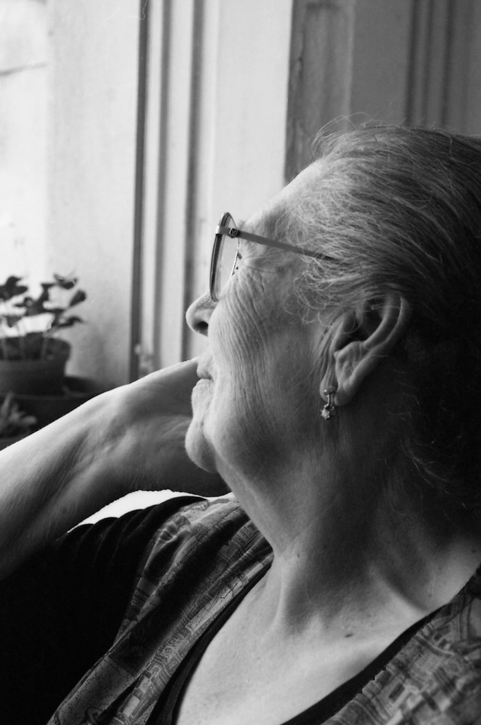 In black and white, an older woman in glasses looks out of a window.