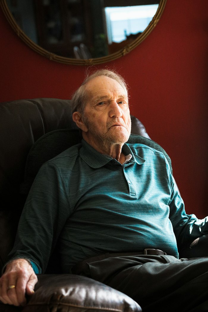 An older man in a green shirt sits back in an armchair against a red wall.