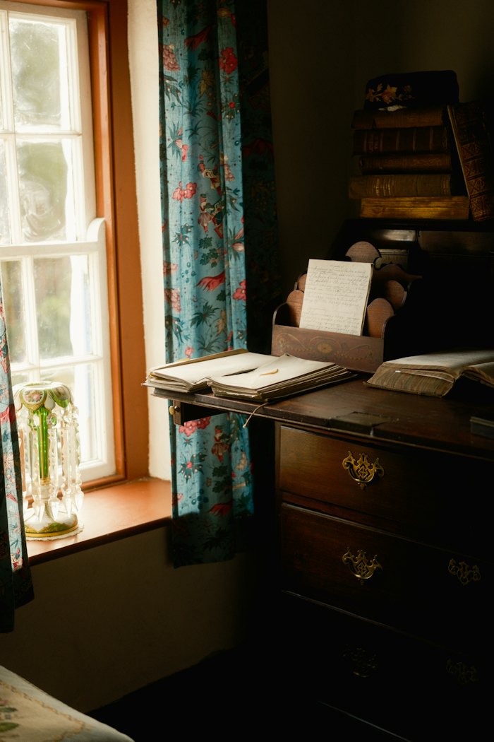 A writing desk with papers beside a sunlit window and patterned curtains.