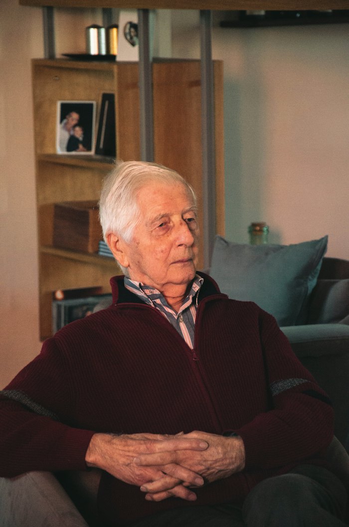 An older man sits in a chair at home with his hands folded, looking to one side.