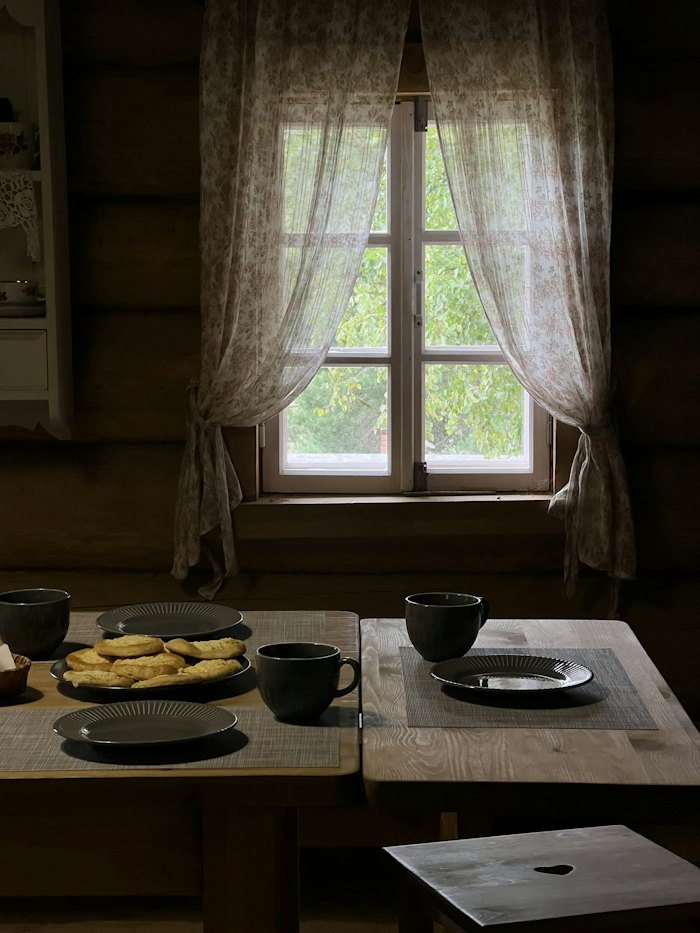 Cups and a plate of pastries on a wooden table beneath a curtained window.